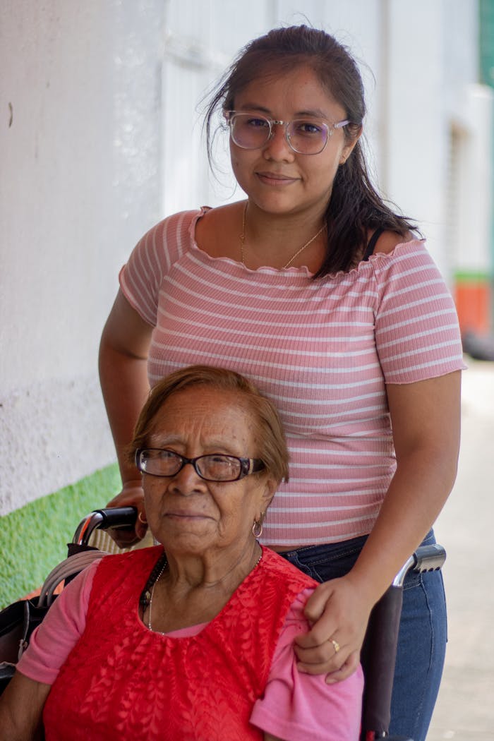 A young woman supports an elderly lady in a wheelchair, showcasing care and compassion outdoors.